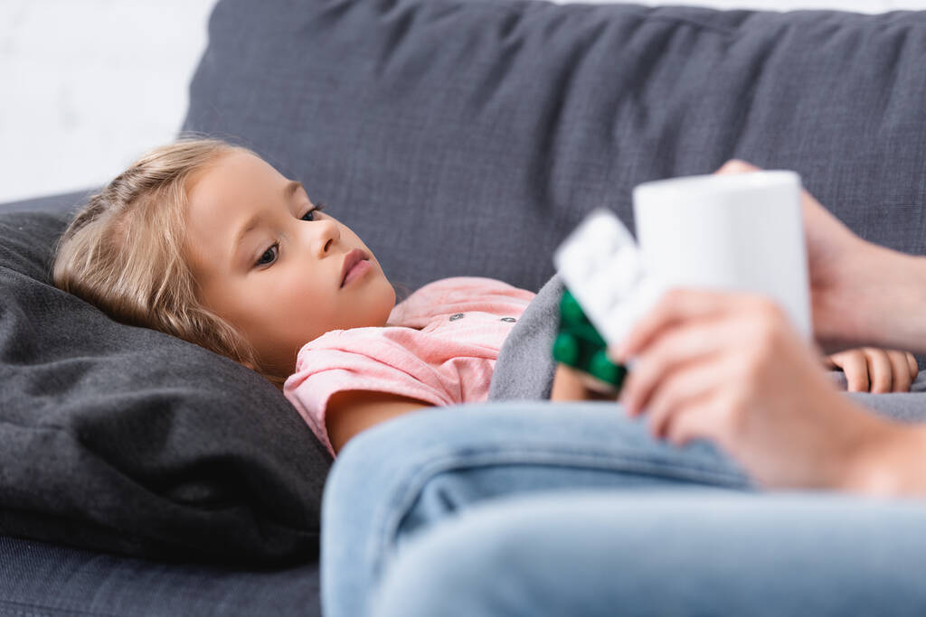Enfoque selectivo de la mujer sosteniendo taza y pastillas cerca del niño enfermo en el sofá  - Foto, Imagen