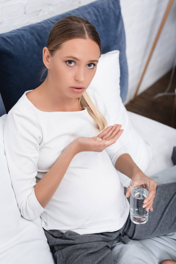 Νεαρή έγκυος γυναίκα κοιτάζοντας κάμερα, ενώ κρατώντας χάπια και ένα ποτήρι νερό  - Φωτογραφία, εικόνα