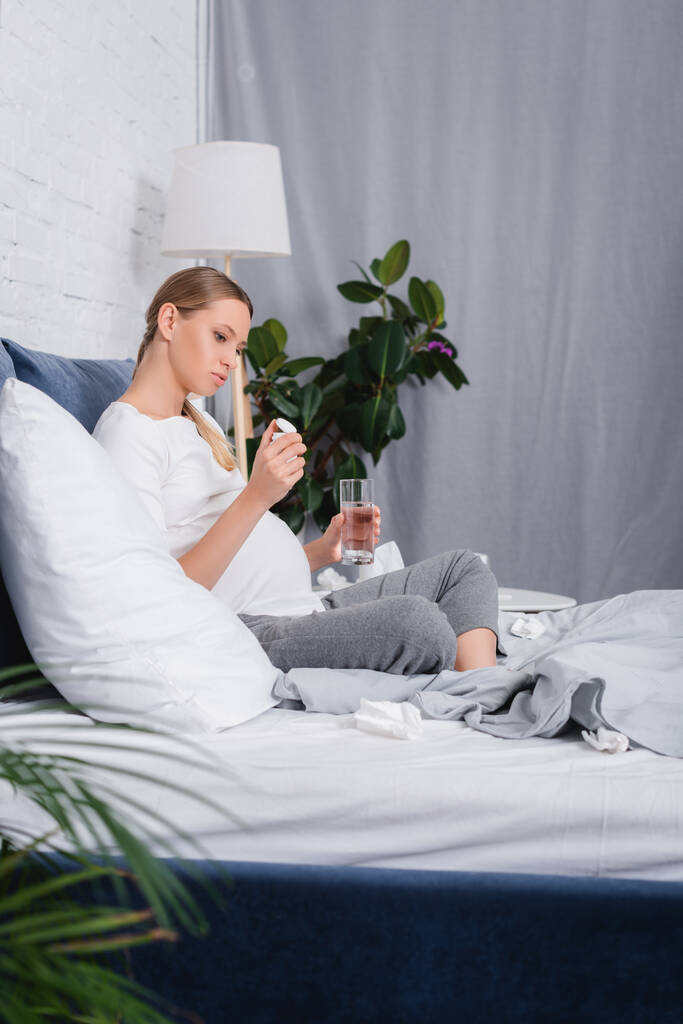 Enfoque selectivo de la mujer embarazada mirando frasco con pastillas y sosteniendo vaso de agua cerca de servilletas en la cama  - Foto, Imagen