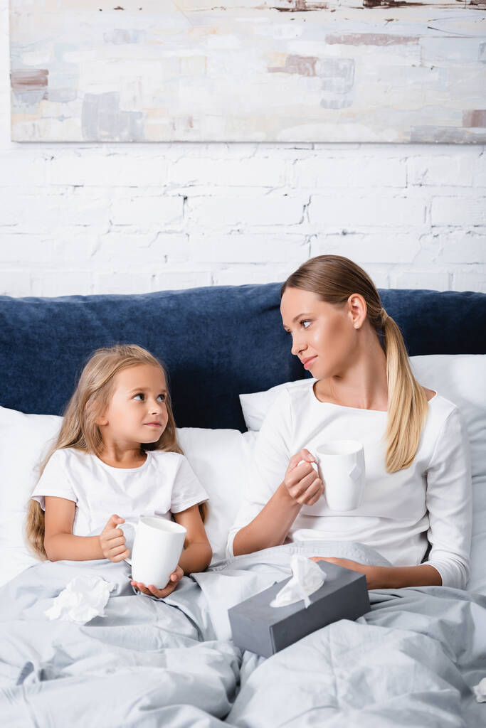 Szelektív fókusz az anya és a gyerek néz egymásra, miközben a csészék közelében szalvéták az ágyon  - Fotó, kép