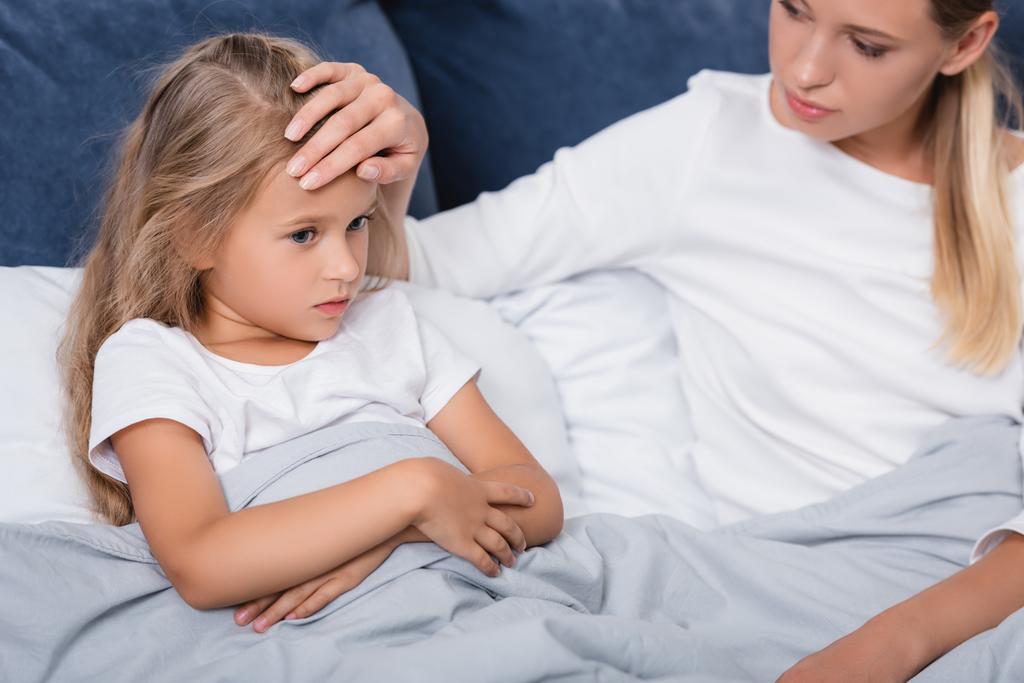 Concentration sélective de la femme touchant le front de la fille malade alors qu'elle est assise sur le lit  - Photo, image
