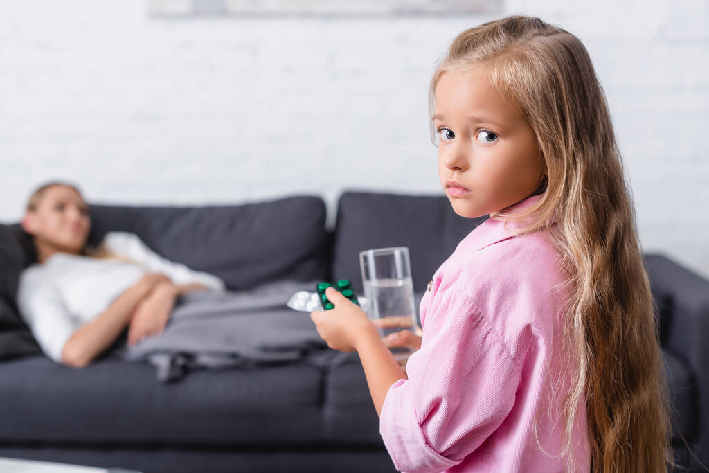 Foco seletivo da menina olhando para a câmera enquanto segurando pílulas um copo de água perto de mulher doente em casa  - Foto, Imagem