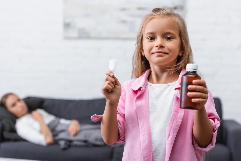 Focus selettivo della ragazza che tiene cucchiaio e bottiglia di sciroppo vicino alla madre malata in soggiorno  - Foto, immagini