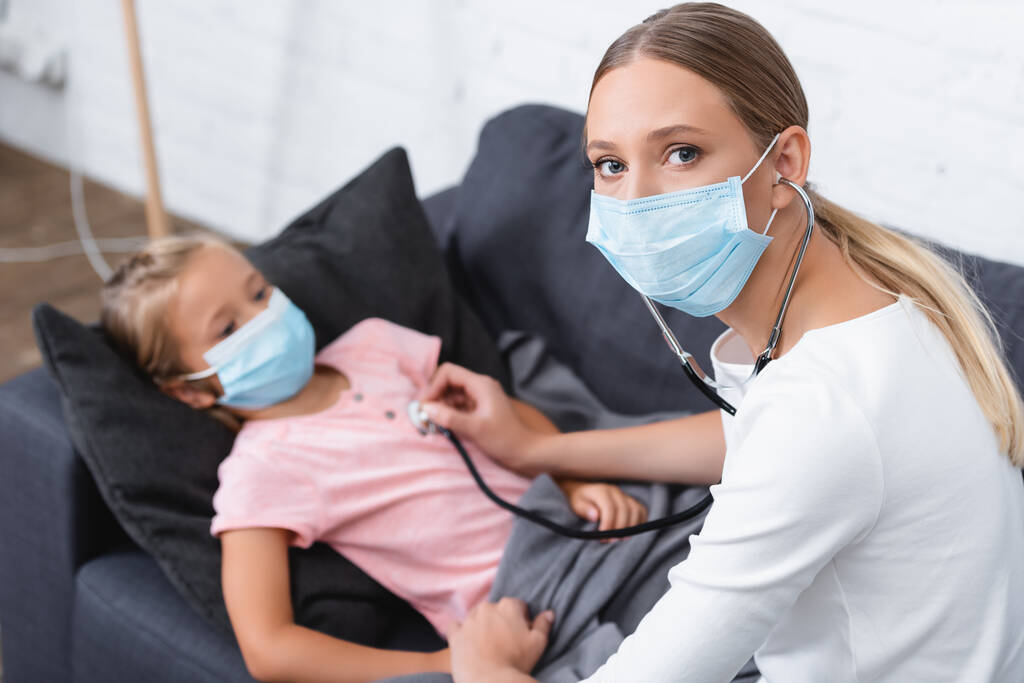 Szelektív fókusz nő orvosi maszk és sztetoszkóp ül gyermek közelében otthon  - Fotó, kép
