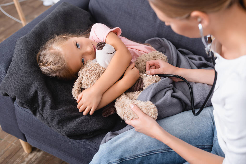Selectieve focus van kind met teddybeer kijkend naar camera bij moeder in stethoscoop - Foto, afbeelding