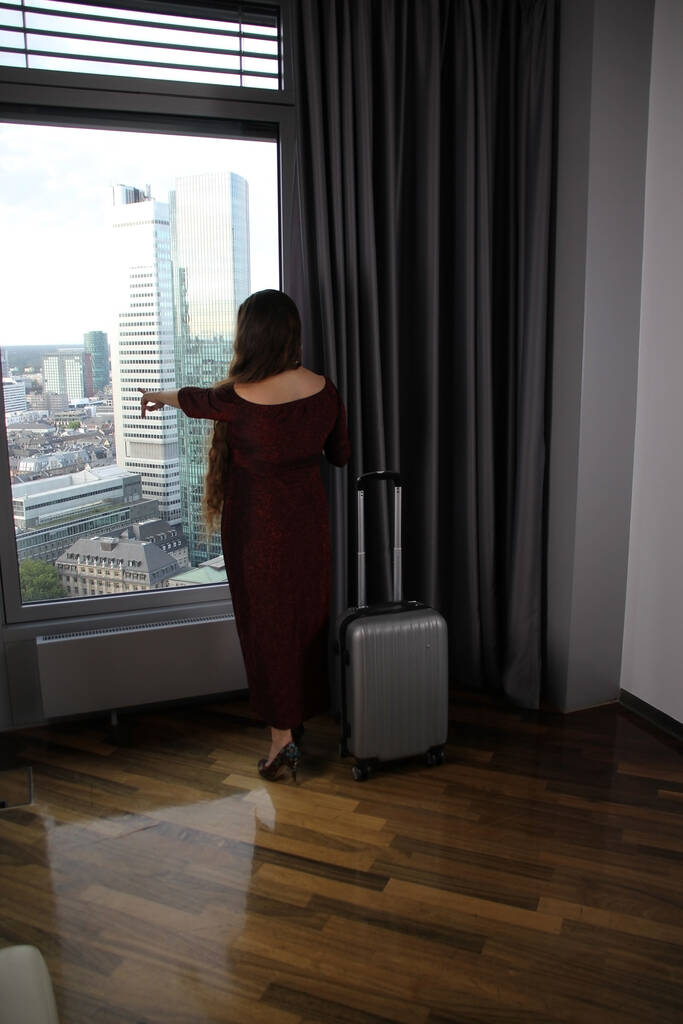 junge Frau in schönem langen Kleid steht am Fenster im Hotel und blickt auf die Stadt, neben den Vorhängen steht ein Koffer, das Konzept einer Geschäftsreise, Reise, Tourismus - Foto, Bild