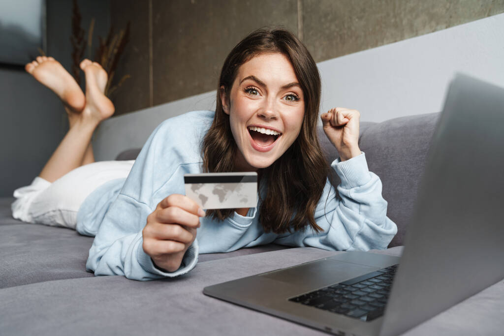 Imagen de la mujer encantada haciendo gesto ganador y la celebración de la tarjeta de crédito mientras trabaja con el ordenador portátil en el sofá - Foto, Imagen