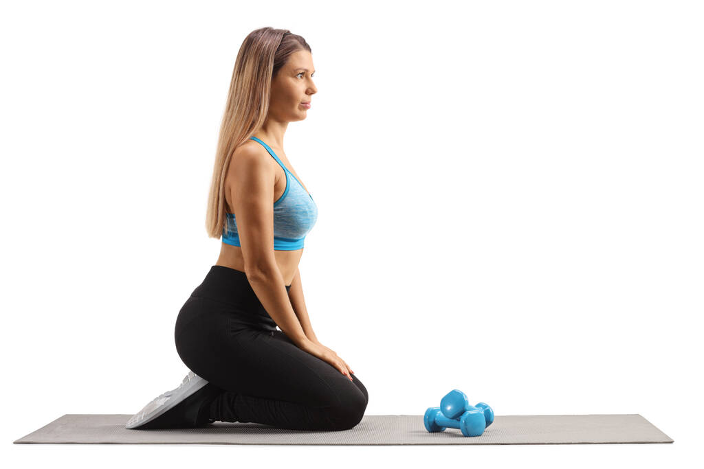 Молодая женщина, стоящая на коленях на коврике для упражнений и готовясь к упражнениям с небольшими весами изолированы на белом фоне - Фото, изображение