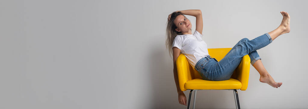 Belle jeune femme est assise ou allongée sur une chaise jaune avec les jambes levées et regardant avec arrogance la caméra, prise de vue en studio, espace de copie, bannière - Photo, image