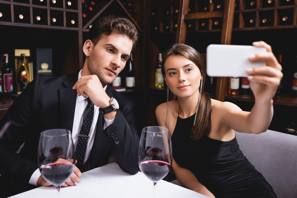 Selectieve focus van elegante vrouw die selfie in de buurt van vriend en glas wijn in restaurant  - Foto, afbeelding