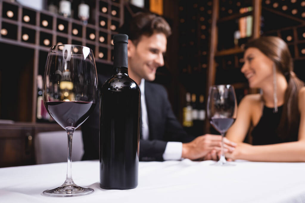 Selektywne skupienie butelki i kieliszka wina na stole w pobliżu młodych par trzymających się za ręce podczas randek w restauracji  - Zdjęcie, obraz