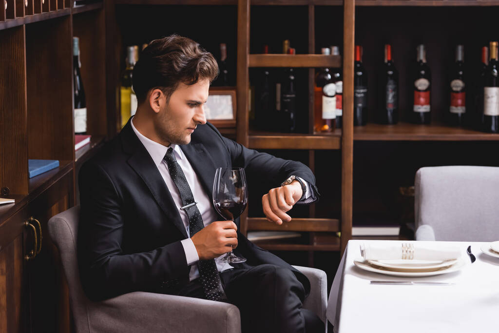 Enfoque selectivo del hombre de traje mirando el reloj de pulsera y sosteniendo una copa de vino en el restaurante  - Foto, imagen