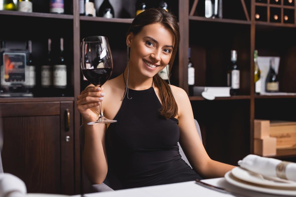 Επιλεκτική εστίαση της κομψής γυναίκας κοιτάζοντας κάμερα, ενώ κρατώντας ένα ποτήρι κρασί στο εστιατόριο  - Φωτογραφία, εικόνα