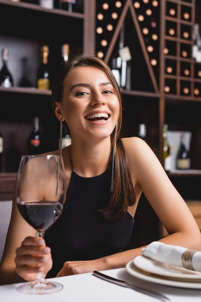 Selectieve focus van opgewonden vrouw die glas wijn vasthoudt terwijl ze naar de camera kijkt aan tafel in restaurant  - Foto, afbeelding