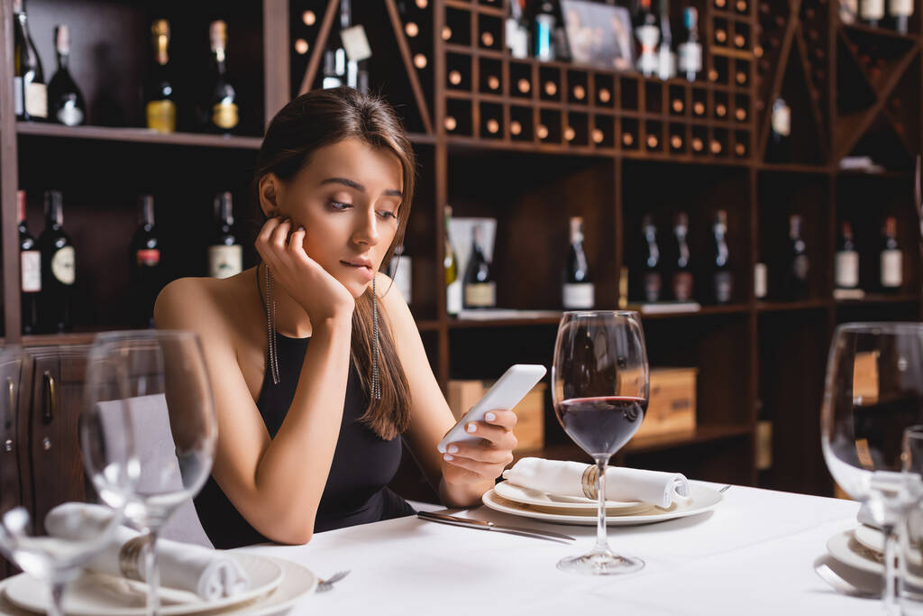 Селективный фокус молодой женщины с помощью смартфона, сидя возле бокала вина в ресторане  - Фото, изображение