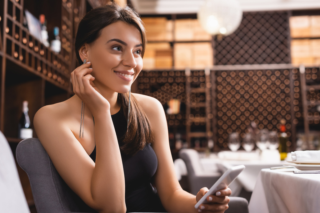 Επιλεκτική εστίαση της κομψής γυναίκας κοιτάζοντας μακριά, ενώ κρατώντας smartphone στο εστιατόριο  - Φωτογραφία, εικόνα