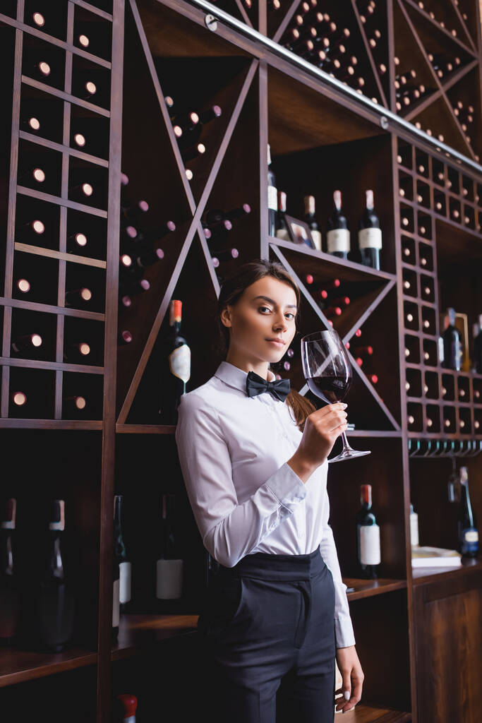 Junger Sommelier mit einem Glas Wein blickt in die Kamera in der Nähe von Regalen mit Flaschen im Restaurant  - Foto, Bild