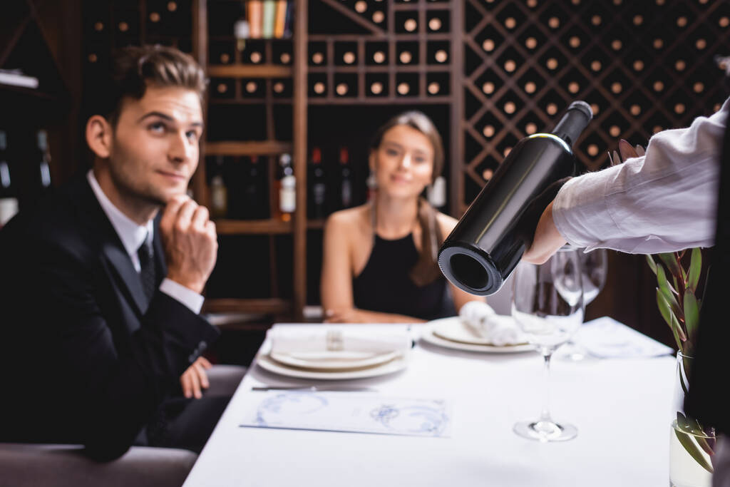 Επιλεκτική εστίαση του sommelier κρατώντας μπουκάλι κρασί κοντά κομψό ζευγάρι στο εστιατόριο  - Φωτογραφία, εικόνα