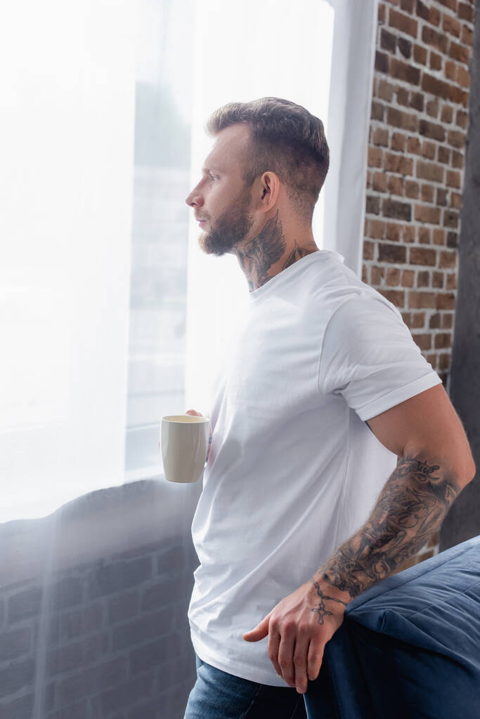 вид сбоку на молодого человека с татуировкой, который смотрит в сторону, стоя у окна с чашкой чая - Фото, изображение