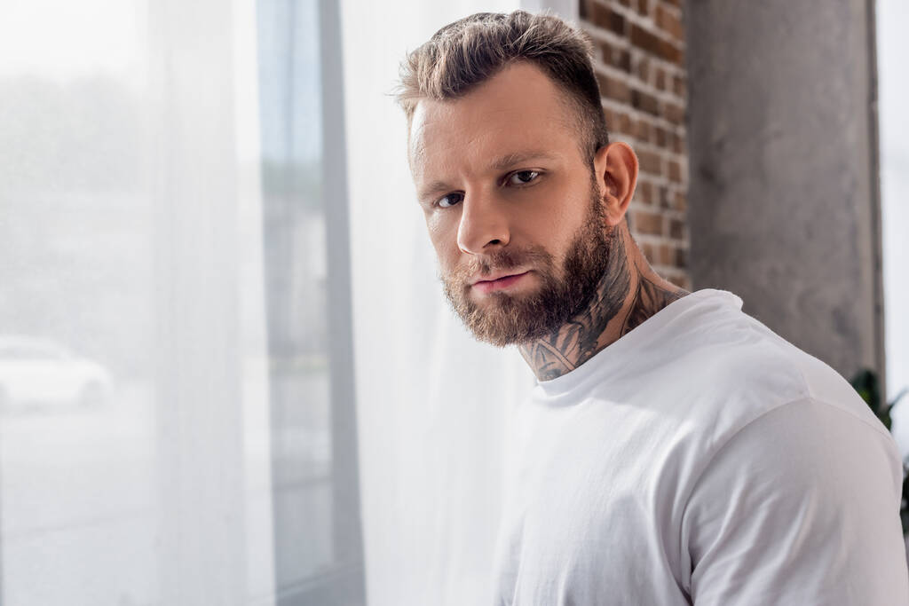bearded, tattooed man in white t-shirt standing near window and looking at camera - Photo, Image