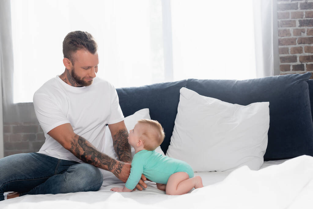 giovane uomo in t-shirt bianca e jeans toccare figlio neonato strisciare sul letto in pagliaccetto bambino - Foto, immagini