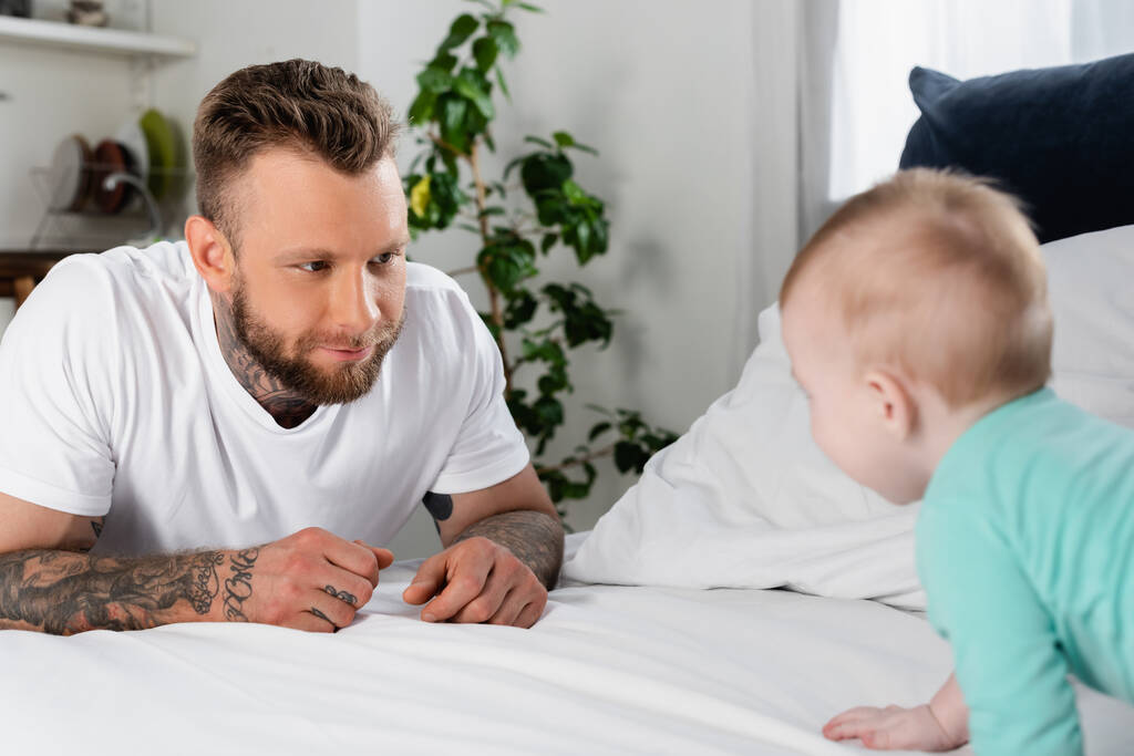 вибірковий фокус татуйованого чоловіка, дивлячись на дитячу повзану на ліжку
 - Фото, зображення
