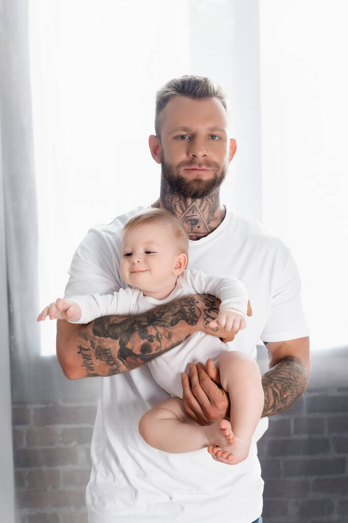 joven hombre tatuado mirando a la cámara mientras sostiene alegre bebé hijo en bebé mameluco - Foto, Imagen