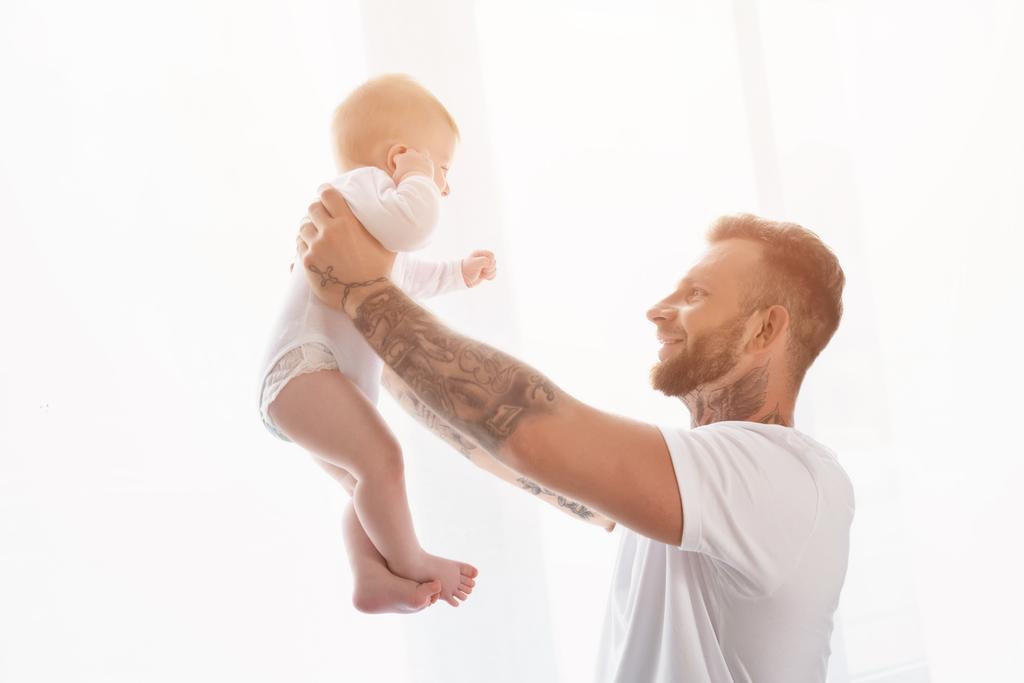 radosny wytatuowany mężczyzna w białej koszulce trzymający niemowlaka w wyciągniętych dłoniach - Zdjęcie, obraz
