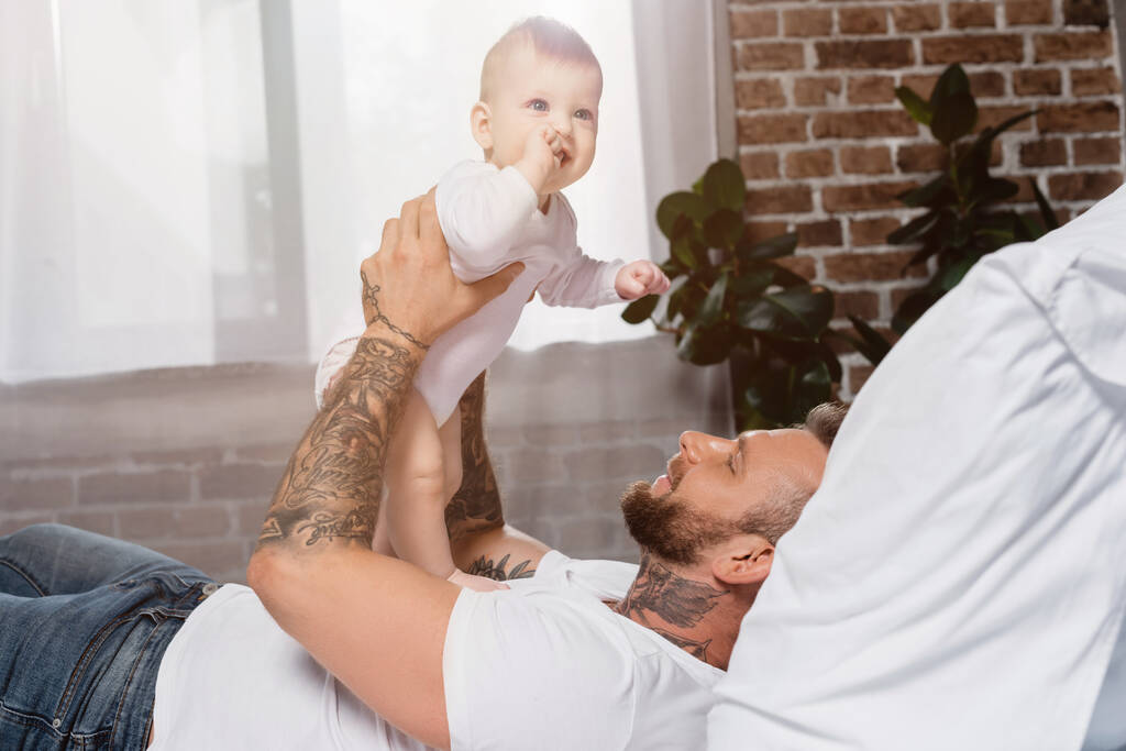 enfoque selectivo de joven hombre tatuado sosteniendo excitado bebé niño mientras está acostado en la cama - Foto, imagen