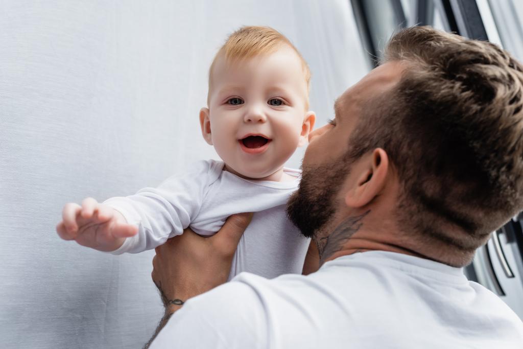 избирательный фокус молодого отца, держащего взволнованного младенца, смотрящего в камеру с открытым ртом - Фото, изображение