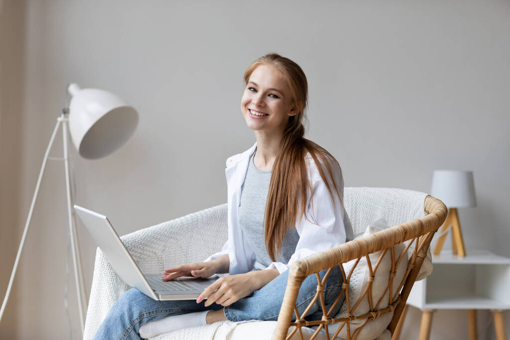 自宅の椅子に座ってノートパソコンを持つ喜びの女性。遠くでのコミュニケーション. - 写真・画像