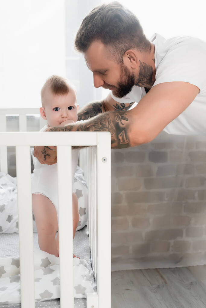 Tätowierter Mann unterstützt Säugling, der im Kinderbett steht und die Hand im Mund hält - Foto, Bild