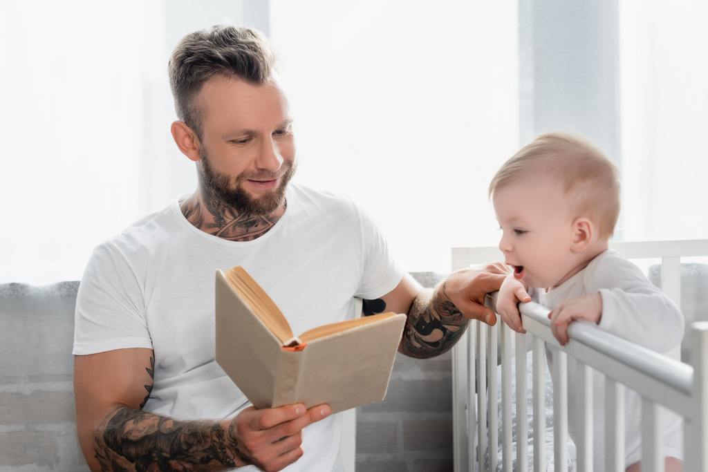 若い入れ墨の男は本を読んで興奮した幼児の男の子にベビーベッドに立って - 写真・画像
