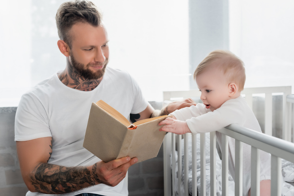 Bebek beşikte duruyor ve genç babanın elindeki kitaba dokunuyor. - Fotoğraf, Görsel