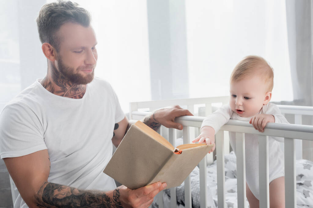 joven padre tatuado lectura libro a bebé niño de pie en cuna - Foto, imagen