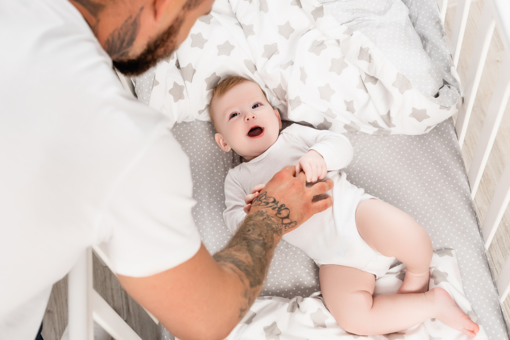 幼児に触れる入れ墨の男の選択的な焦点は口を開けてベビーベッドに横たわっています - 写真・画像