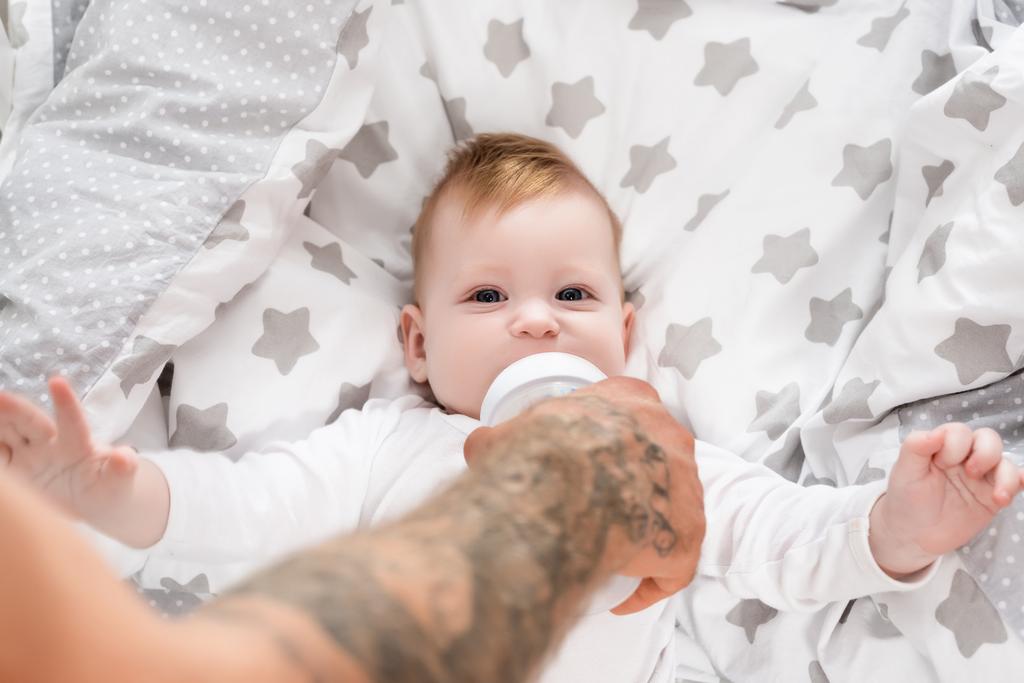 szelektív fókusz tetovált férfi etetés csecsemő fia feküdt bölcsőben cumisüvegből - Fotó, kép