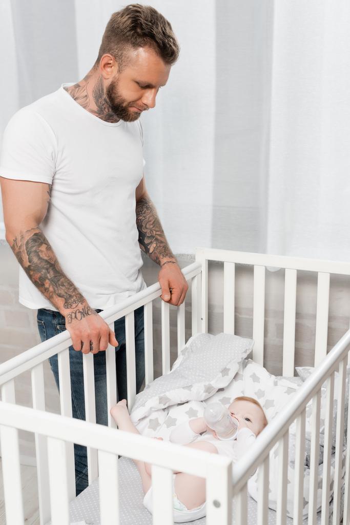 молодой татуированный мужчина смотрит на младенца лежащего в кроватке с детской бутылочкой - Фото, изображение
