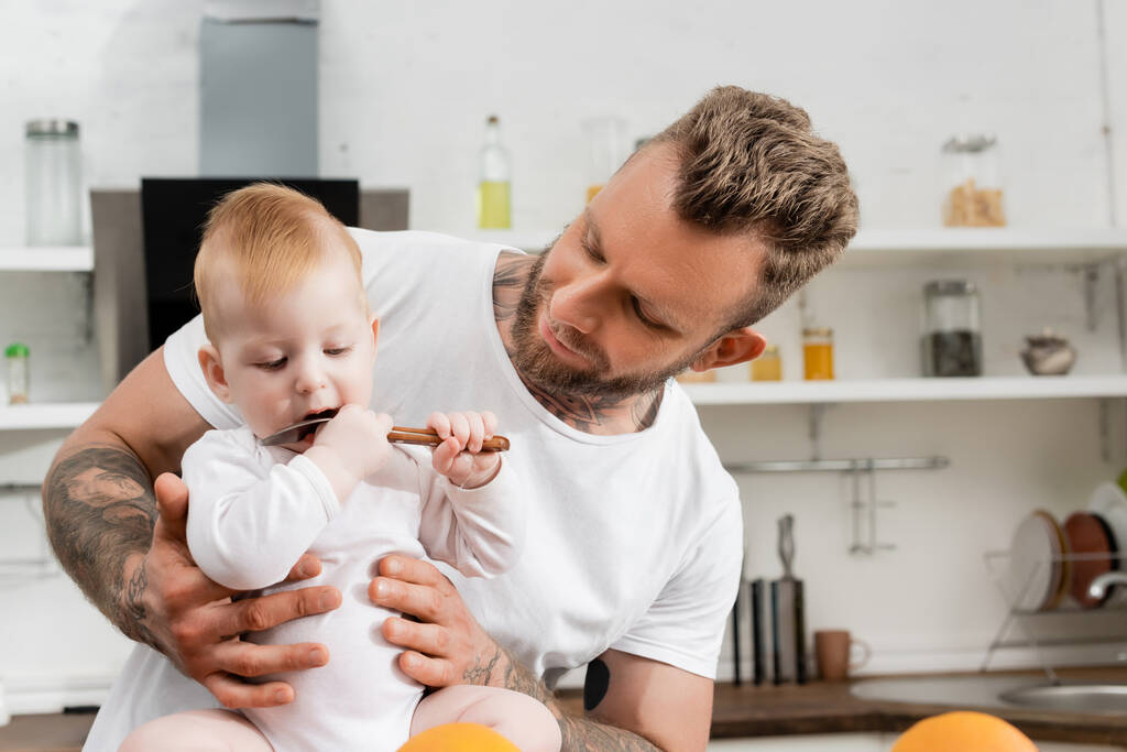 foco seletivo do bebê menino lambendo colher perto do jovem pai na cozinha - Foto, Imagem