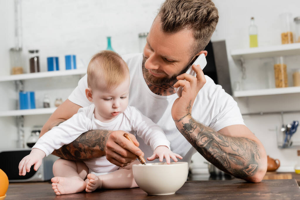 young father talking on smartphone while mixing breakfast in bowl near infant son - Photo, Image