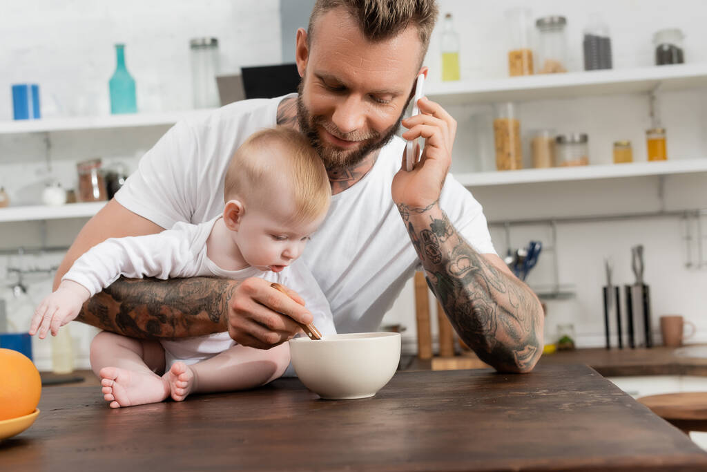 młody brodaty człowiek mówi na smartfonie w pobliżu chłopca i miski ze śniadaniem w kuchni - Zdjęcie, obraz