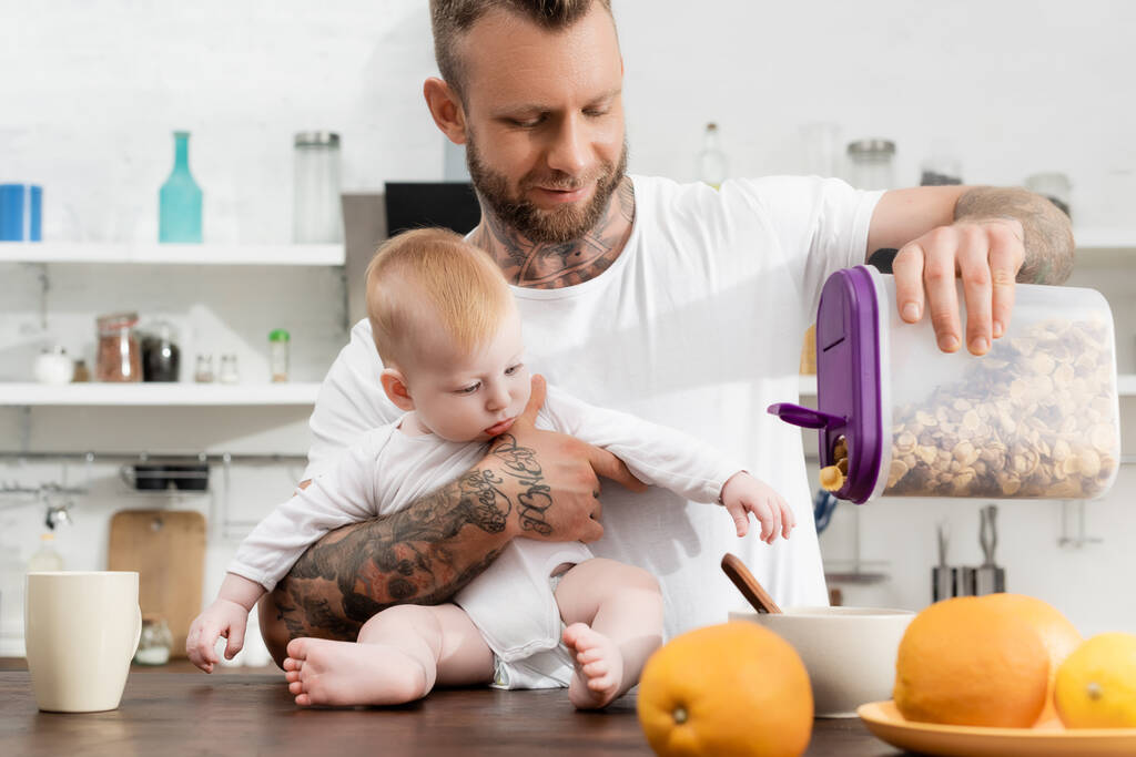 giovane uomo tatuato versando cornflakes in ciotola durante la colazione con il piccolo figlio - Foto, immagini