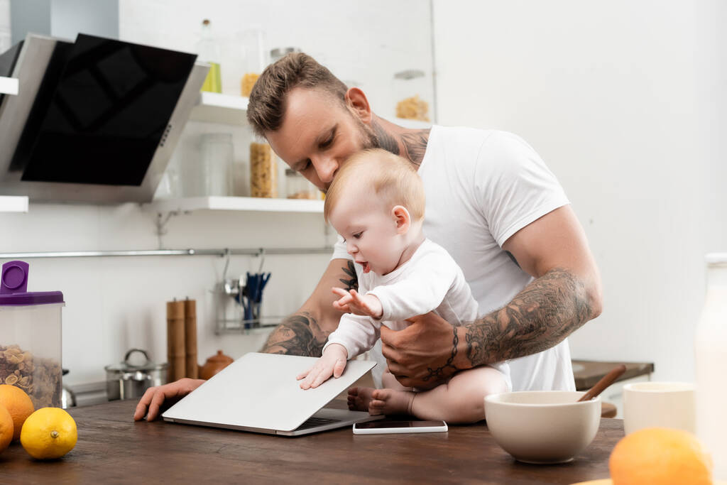 bébé garçon toucher ordinateur portable tout en étant assis sur la table de cuisine près de père tatoué - Photo, image
