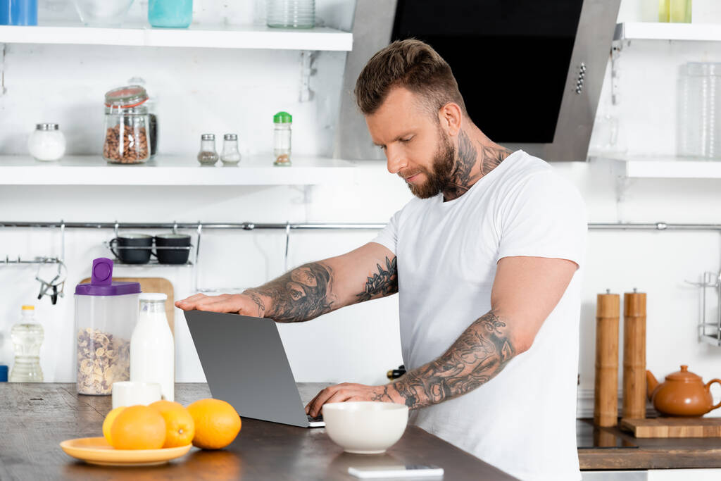 Tätowierte Freiberuflerin im weißen T-Shirt mit Laptop in der Nähe von Milchflasche, Schüssel und Orangen in der Küche - Foto, Bild