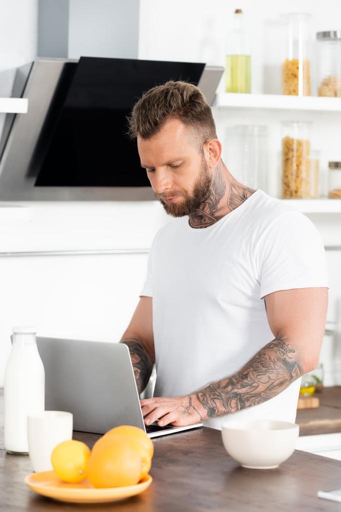 fiatal tetovált szabadúszó segítségével laptop közelében tál, narancs, és üveg tej a konyhában - Fotó, kép