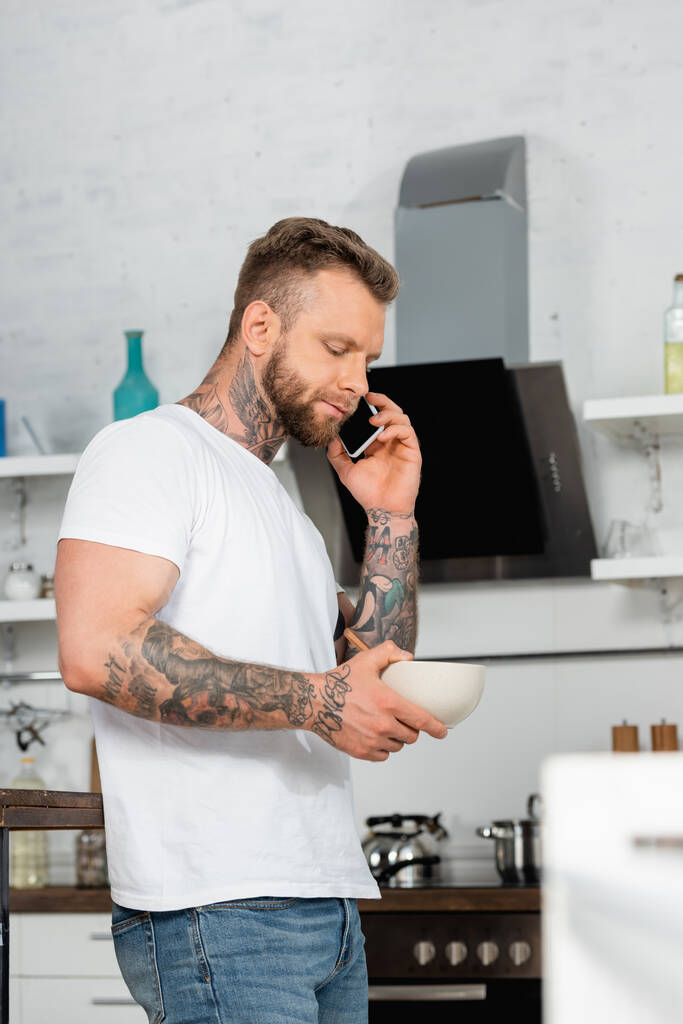 focus selettivo dell'uomo tatuato in t-shirt bianca che parla sul cellulare mentre tiene in mano la ciotola con la colazione - Foto, immagini