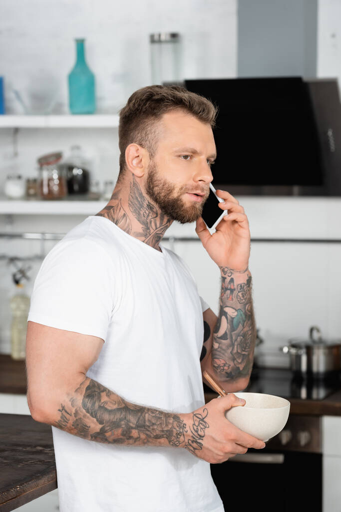 bearded, tattooed man in white t-shirt holding bowl with breakfast while talking on smartphone in kitchen - Photo, Image