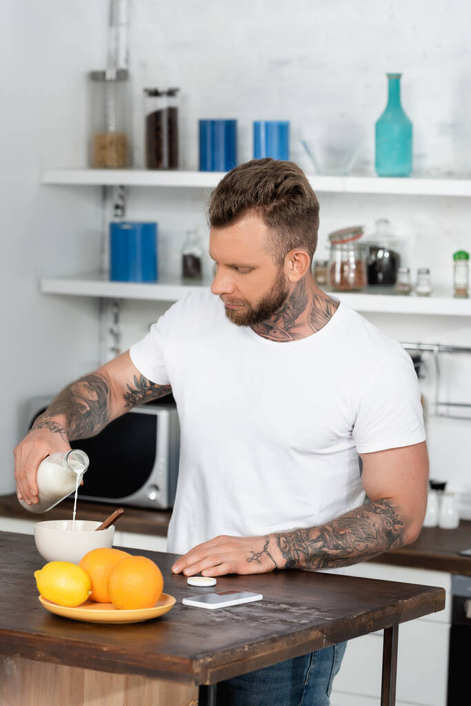 Tätowierter Mann in weißem T-Shirt gießt Milch in Schüssel neben Orangen und Laptop in Küche - Foto, Bild