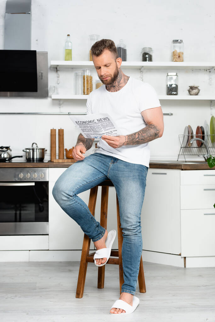 bearded, tattooed man in white t-shirt and jeans reading newspaper while sitting on stool in kitchen - Photo, Image