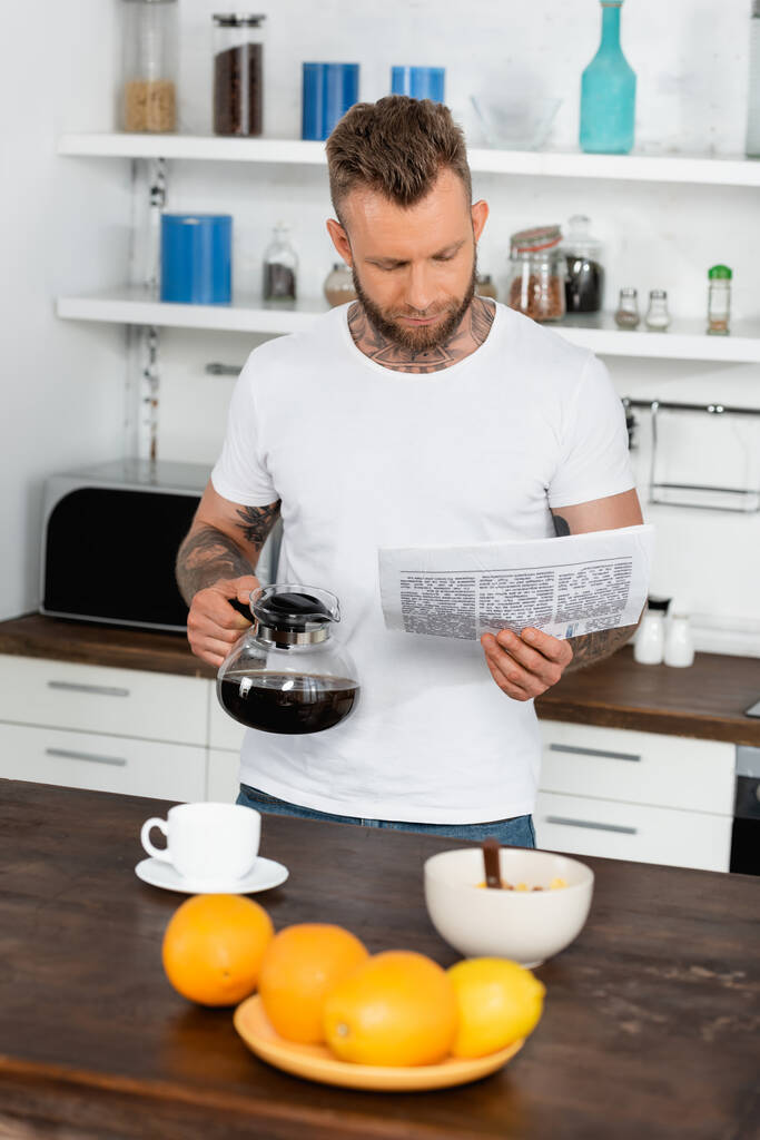 キッチンでコーヒーポットを持ちながら新聞を読む白いTシャツの入れ墨の男の選択的な焦点 - 写真・画像